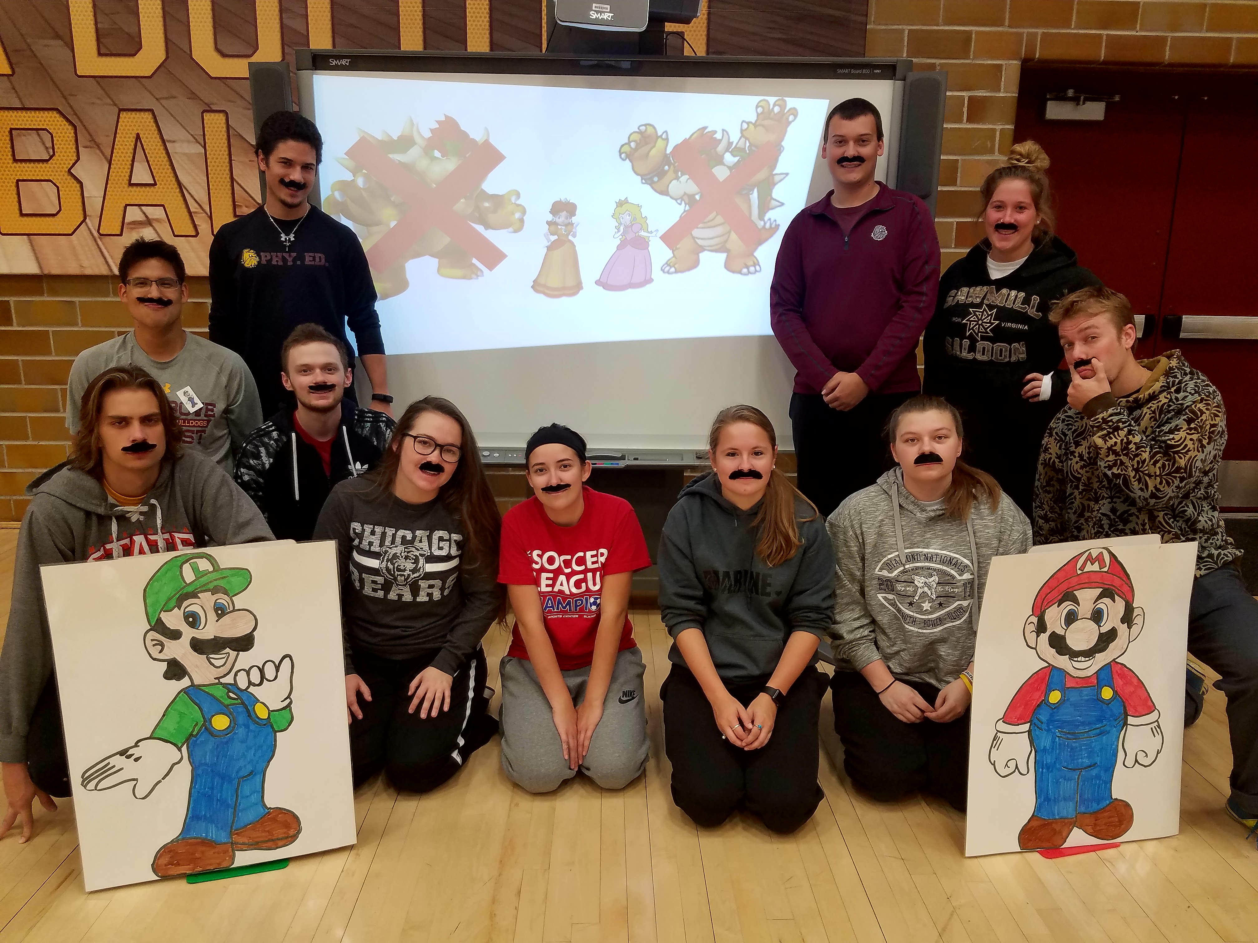 11 students with fake mustaches pose in front a smart board with Mario and Luigi posters