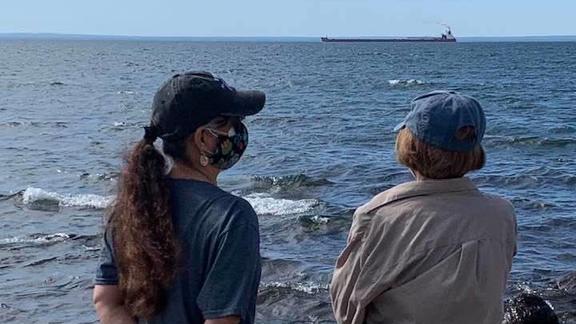 Roxanne Gould and her mother making an offering of asema (tobacco) at Lake Superior with a ship in the distance.
