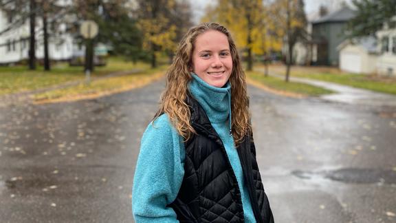 Becca Osborne, a senior at UMD smiling for a photo in the road (wet from rain) with yellow and green trees in the background.