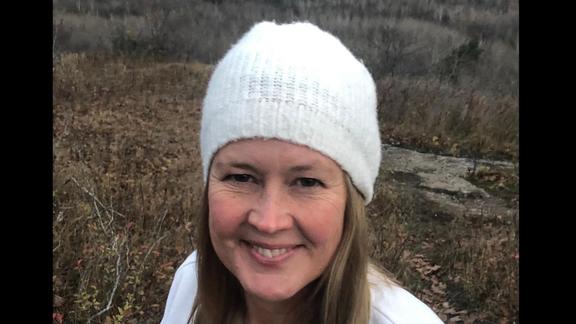 Carina Barker smiling for a photo wearing a white hat with leaves and trees behind her--showing the signs that winter is approaching
