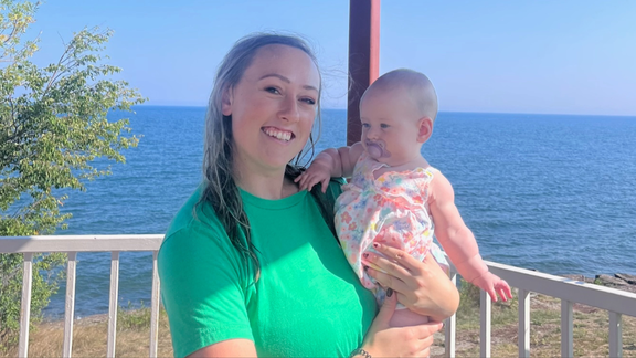 Liz Urbaniak smiling, holding a baby in front of Lake Superior