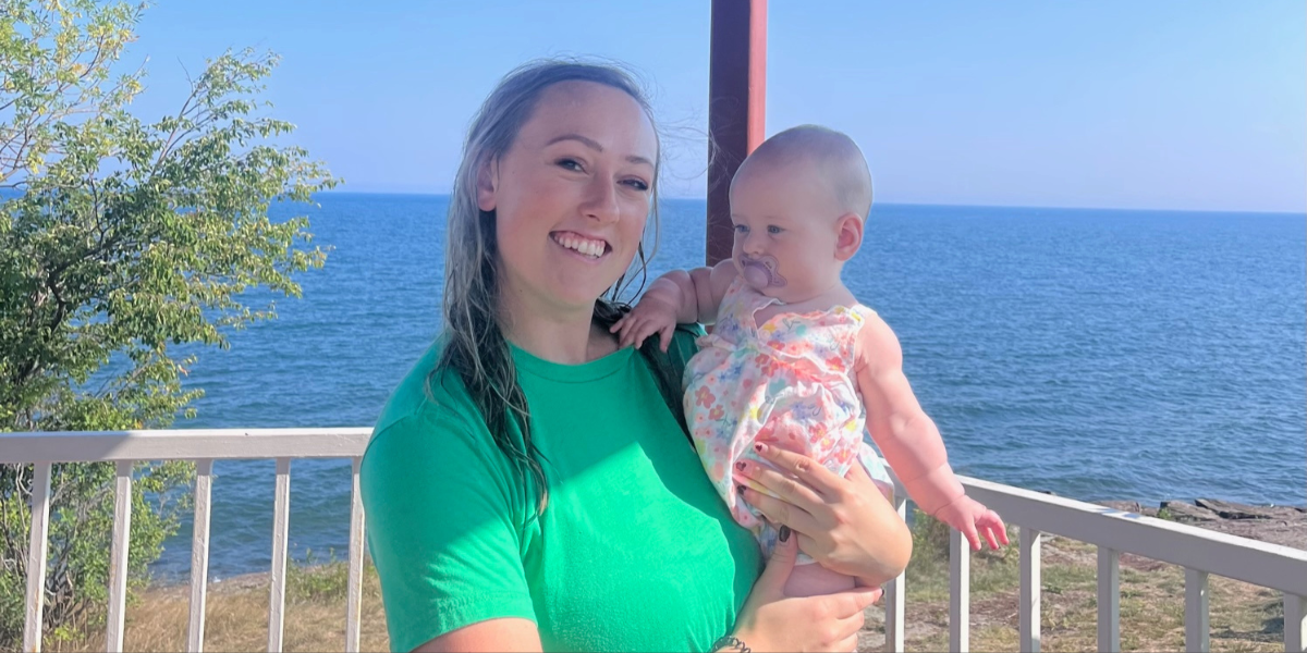 Liz Urbaniak smiling, holding a baby in front of Lake Superior
