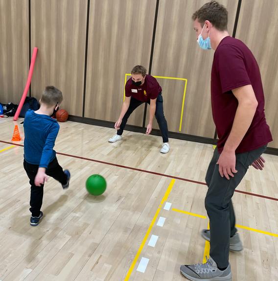 Child kicking a ball towards an instructor in front of a net with another instructor looking on