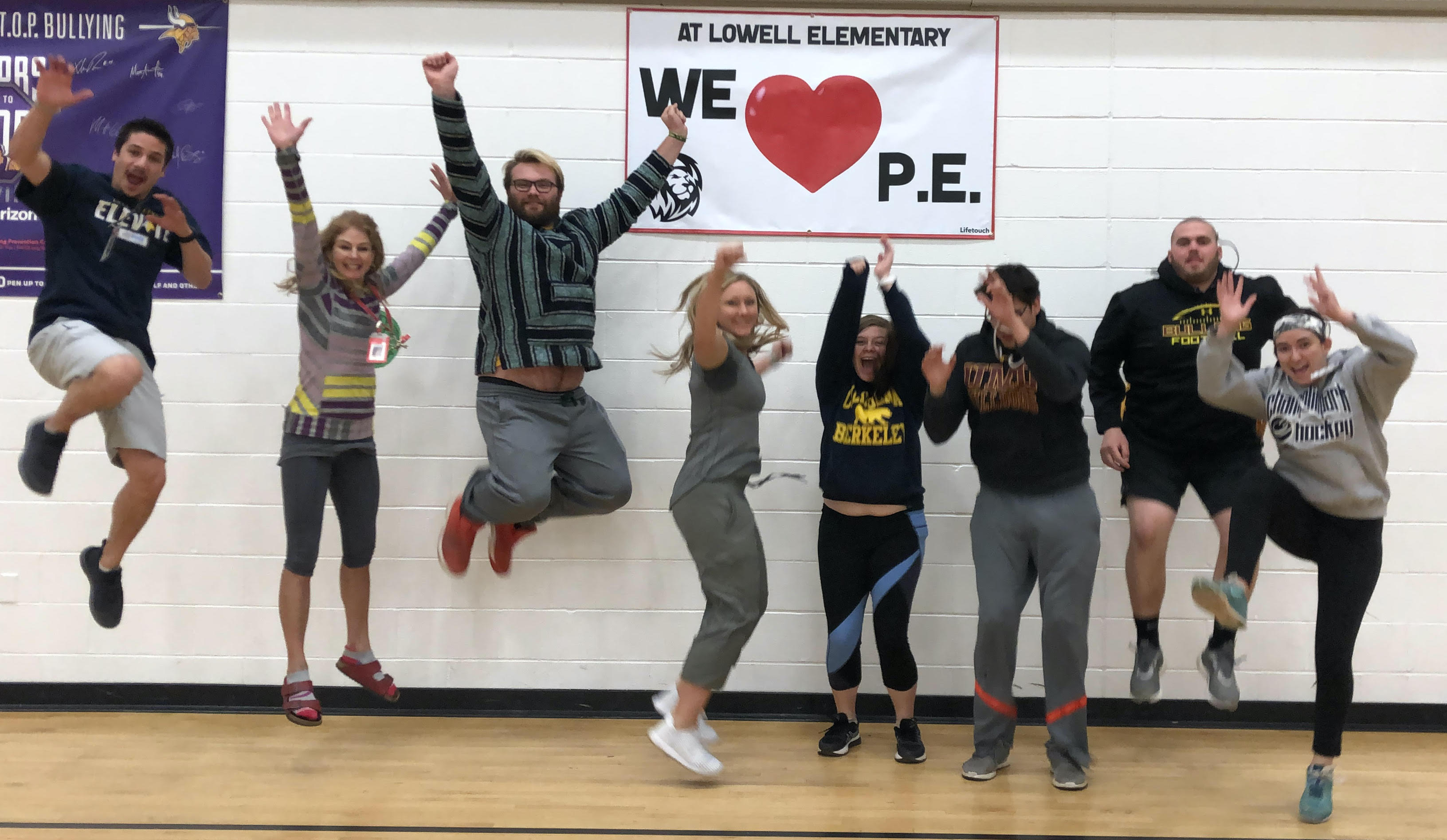 8 adults jumping with excitement for a photo op in a elementary school gymnasium