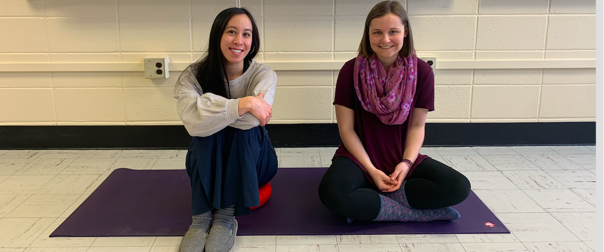 Viann Nguyen-Feng and Kelsey Dietrich, sitting on a yoga mat, smiling