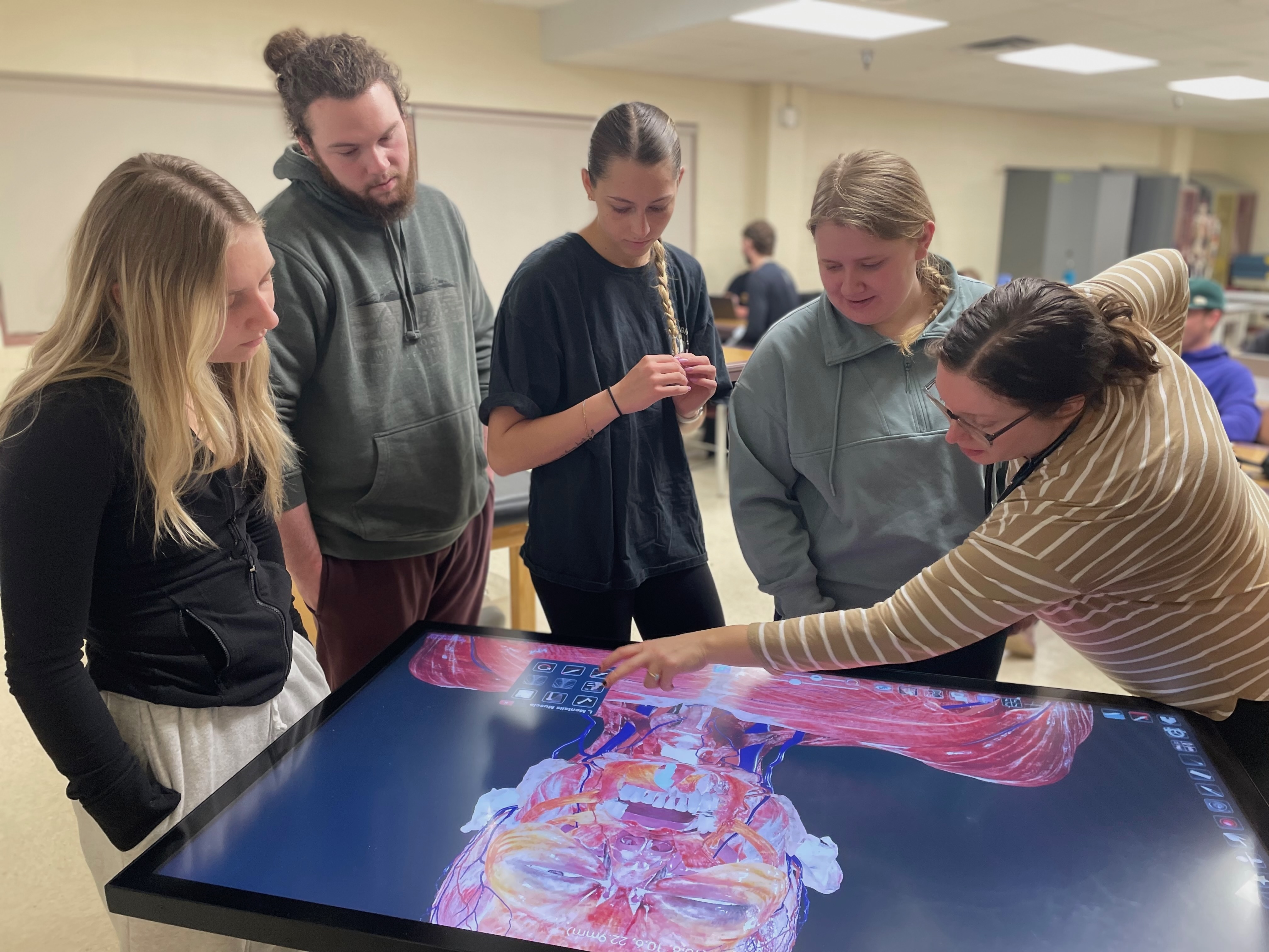 professor pointing to vivid digital display table with human head and shoulders students looking on