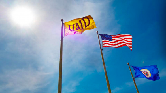 A UMD flag, an American flag, and a MN flag with a blue, sunny sky in the background