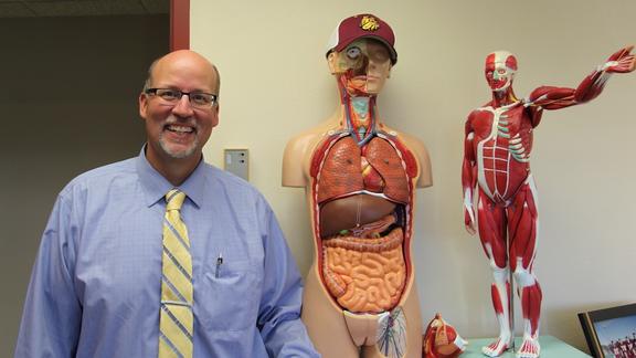 Mike Wendinger wearing a blue shirt and yellow tie and posing next to models of the human body.