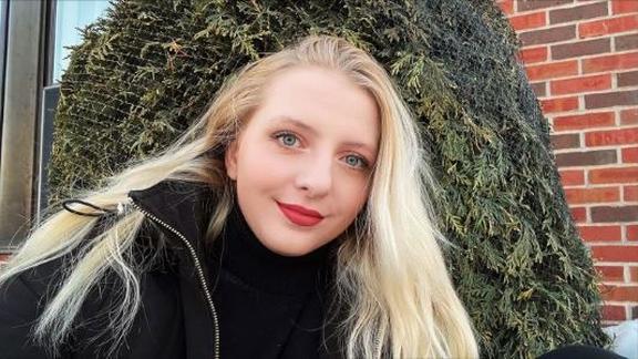 Nicole strumbel in front of brick wall and pine tree. she has bright blond hair, red lipstick, and is wearing a black coat