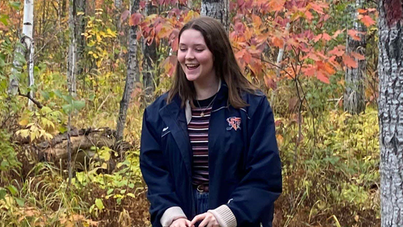 Student worker Molly Baumeister laughing in an autumn colored forest wearing a tan knit sweater, navy blue jacket, and purple striped shirt. 