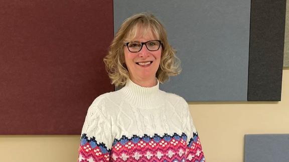 Lynette Carlson is smiling she has a short blonde bob, black square glasses, and a colorful white striped sweater.