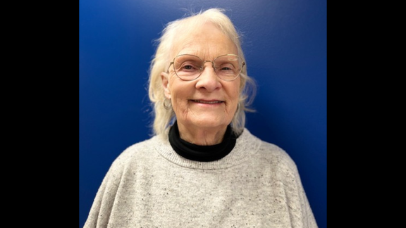 Lynda John, smiling with a beige sweater, glasses and a blue background