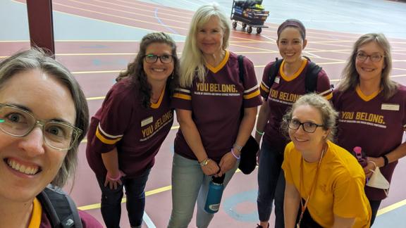 Group photo of advising staff in the field house, smiling and welcoming students and wearing maroon and gold shirts that say "You Belong"