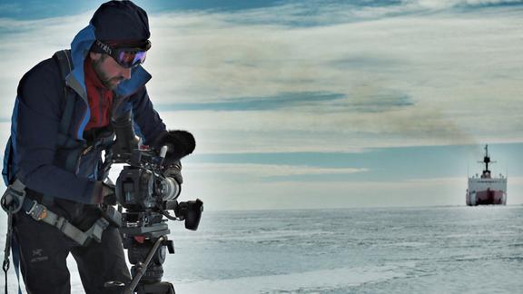 JJ Kelley adjusts his camera in an icy landscape with a ship in the distance
