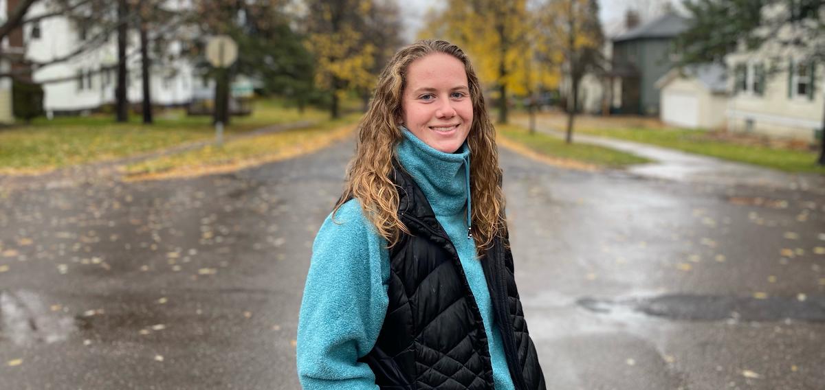 Becca Osborne, a senior at UMD smiling for a photo in the road (wet from rain) with yellow and green trees in the background.
