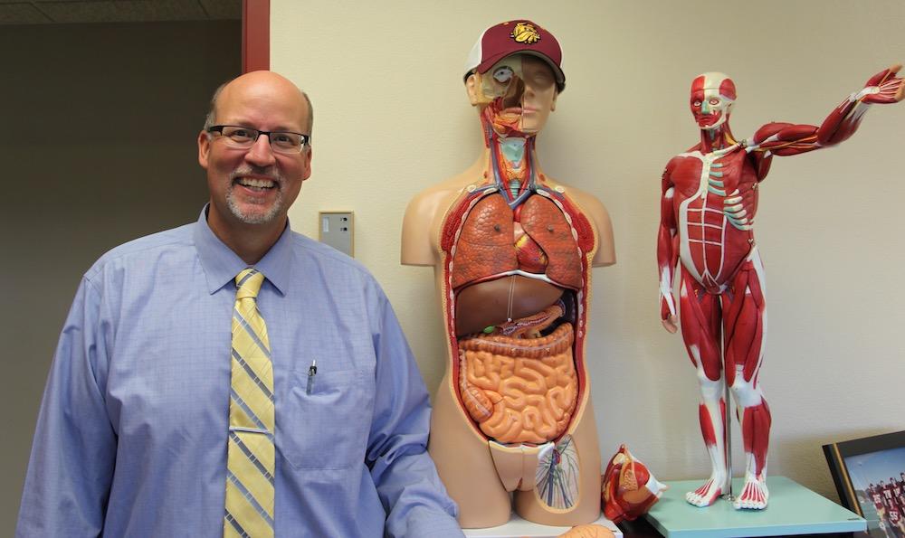 Mike Wendinger wearing a blue shirt and yellow tie and posing next to models of the human body.
