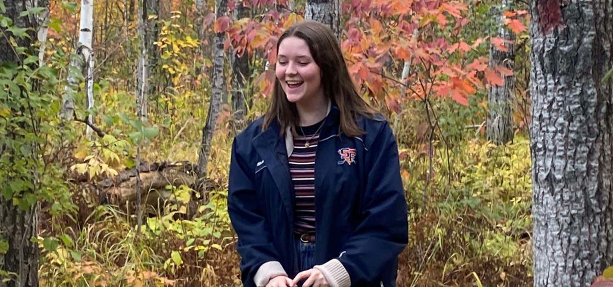 Student worker Molly Baumeister laughing in an autumn colored forest wearing a tan knit sweater, navy blue jacket, and purple striped shirt. 