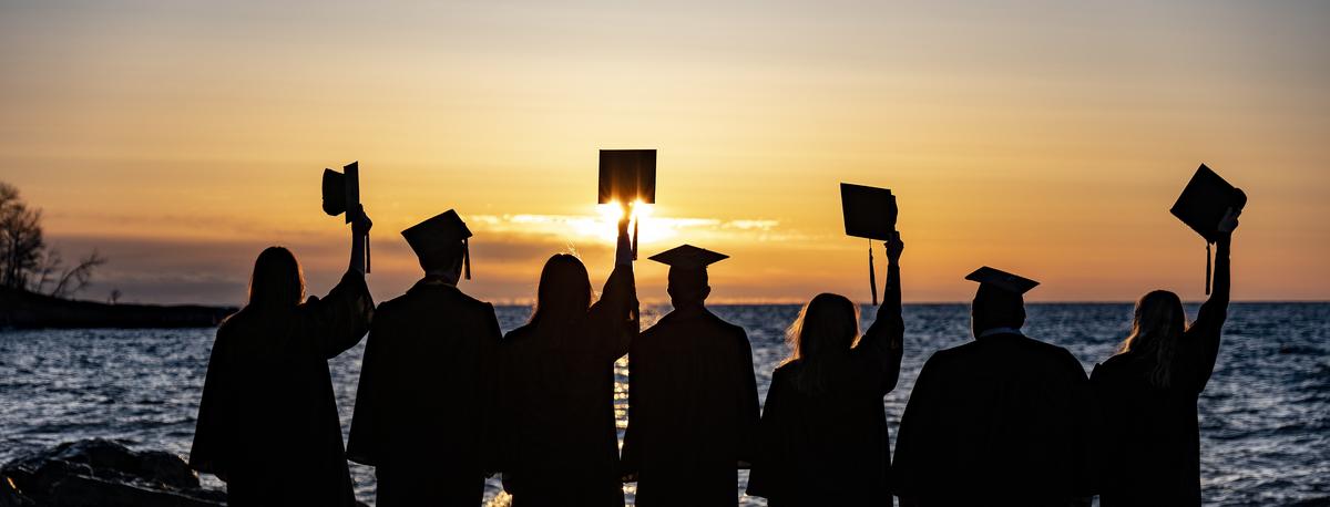 Graduates lifting their caps as they watch the sun rise over lake superior 