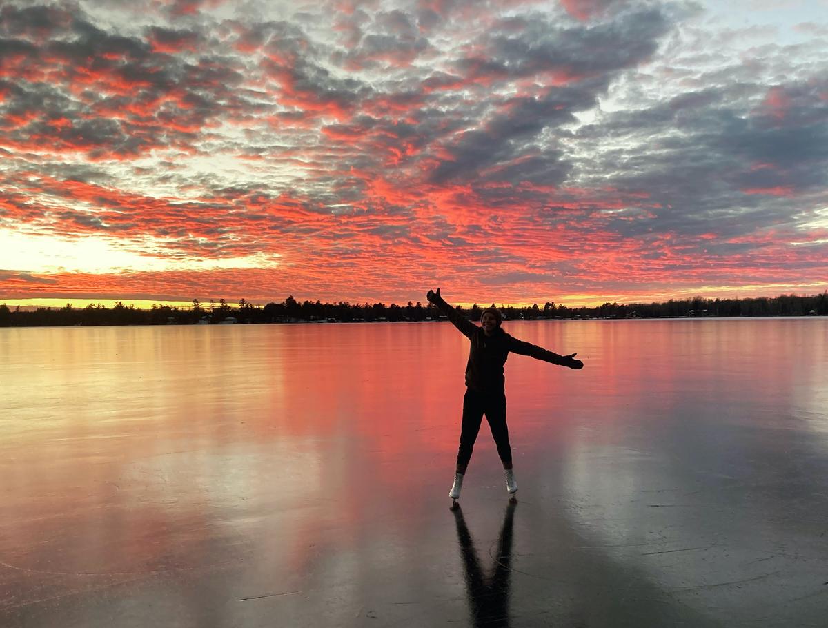 Haley Diem ice skating with arms outstretched and a colorful cloudy sunset backdrop