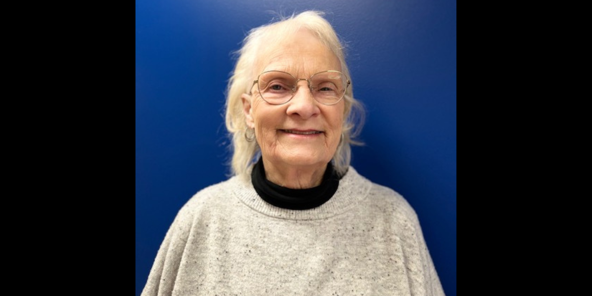 Lynda John, smiling with a beige sweater, glasses and a blue background