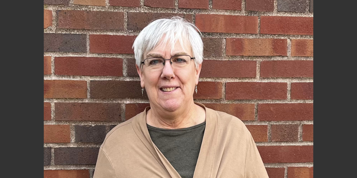 Leslie McFadden, with short white hair and glasses, wearing a tan cardigan and standing against a brick wall