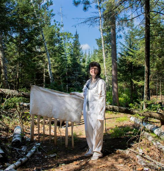 Raina posing in the woods, dressed in white and holding up a white drag cloth used for gathering ticks