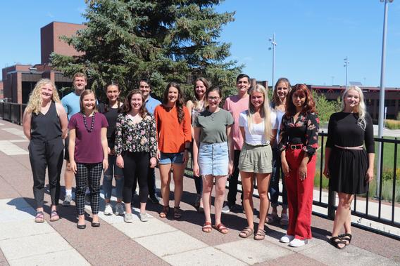 14 Masters of Psychological Science students stand together on a sunny day outdoors.