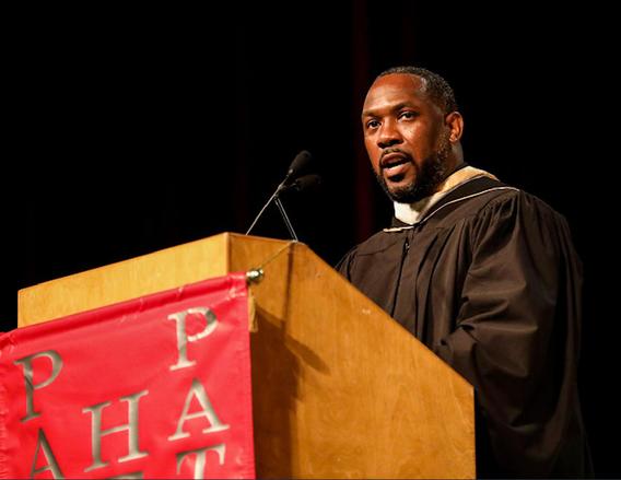 Yusuf Abdullah dressed in graduate robes speaking at a podium
