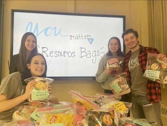4 Students holding up plastic bags in front of a sign that reads "you matter resource bags" the bags have self care items and a motivational note. many more lay on a table in front of them. 
