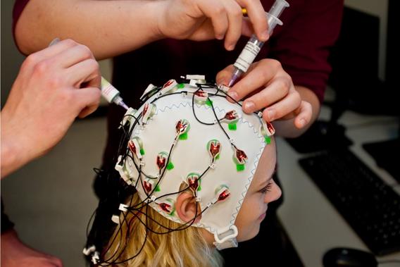 A girl wears a cap with electrodes while two people place syringes with fluid into the sensors.