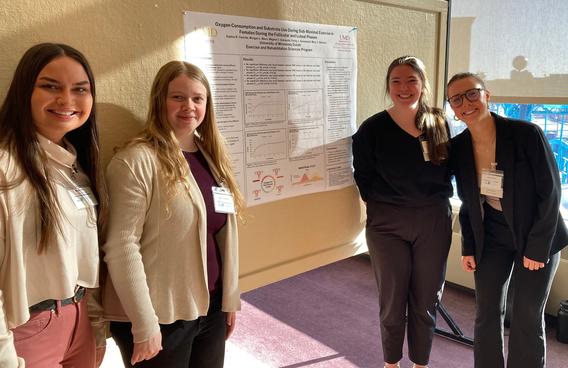 Four students, two on either side of a poster presentation, smiling