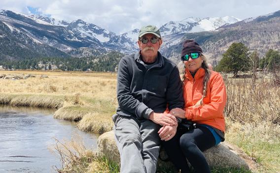 Fay Maas and her husband, wearing hats and sunglasses and sitting next to a mountain lake