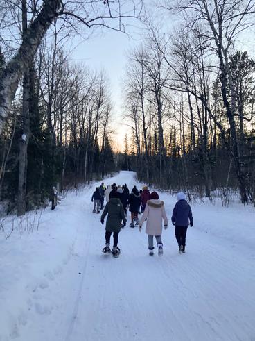 Students snowshoeing down the road with trees and sunset in the background